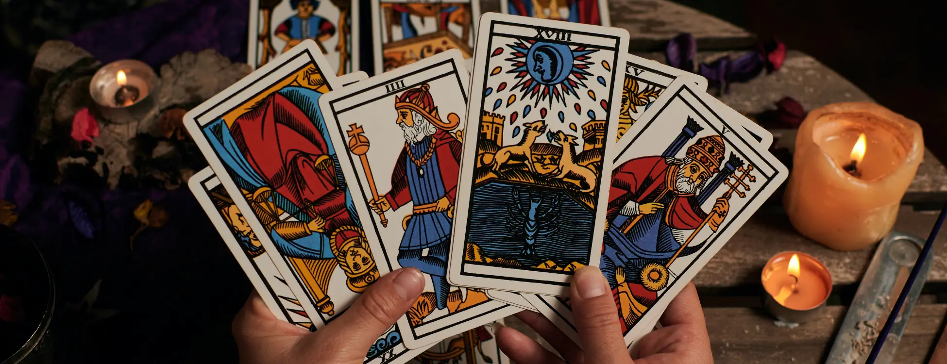 Hands holding illustrated tarot cards beside glowing candles on a wooden table.