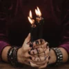 Hands holding black candles against a burgundy blouse, with the face outside the frame.