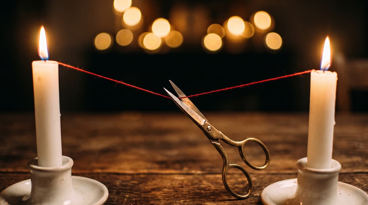 Cord cutting ritual: brass scissors cutting a red thread between two lit candles, a release ritual by PuraTarot