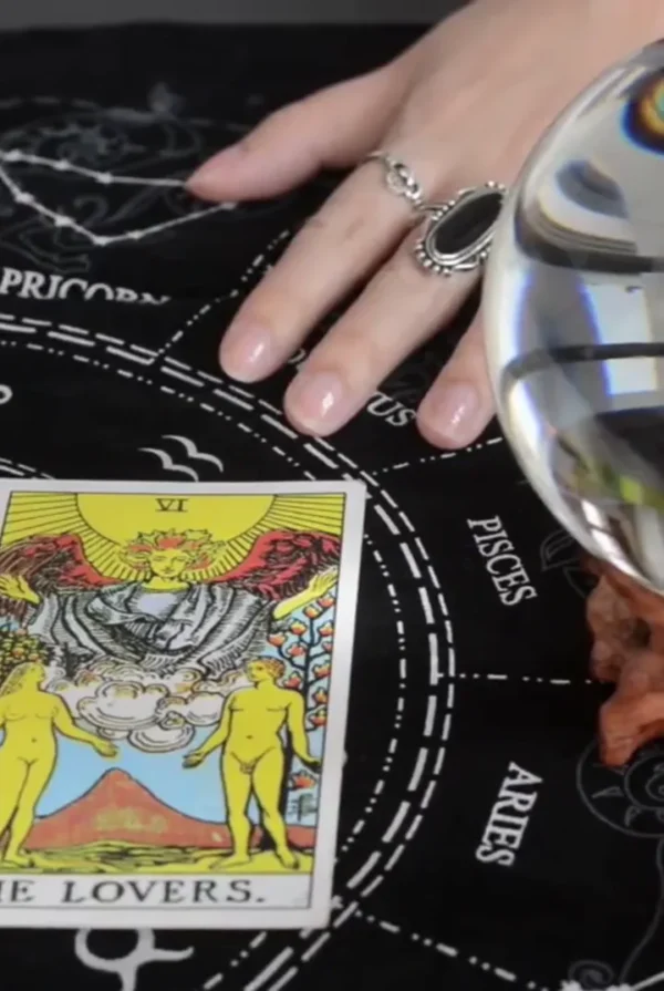 The Lovers tarot card beside a crystal ball and a ringed hand on a black zodiac cloth.